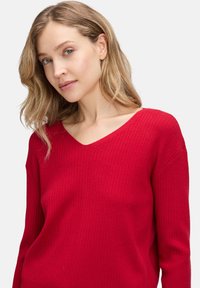 Roter gerippter Strickpullover mit V-Ausschnitt und langen Ärmeln, ausgestattet mit einer strukturierten Oberfläche und einem lässigen Schnitt.