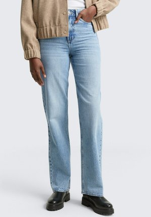 Persona che indossa jeans a gamba dritta blu chiaro, giacca beige, top bianco e stivali neri con suola spessa, in piedi con una mano in tasca.