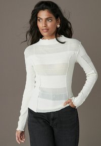 Hvid langærmet top med tekstureret, stribet mønster. Har en høj hals og let rynke ved manchetterne, kombineret med mørke jeans.