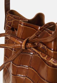 Ruskea nahkainen ämpärilaukku, jossa on krokotiilitekstuuri, nauhakiinnityksellä ja sileällä sisäpinnalla. Koristeet ja ompeleet parantavat muotoilua.