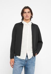 Casaco preto com fecho zip frontal e gola, usado sobre uma camisa clara com botões, combinado com jeans azuis. Tecido suave, corte descontraído, design minimalista.