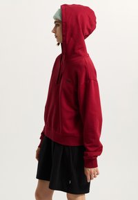 Red hoodie with a front pocket and drawstring hood, paired with a black skirt. The hoodie features ribbed cuffs and a subtle logo.