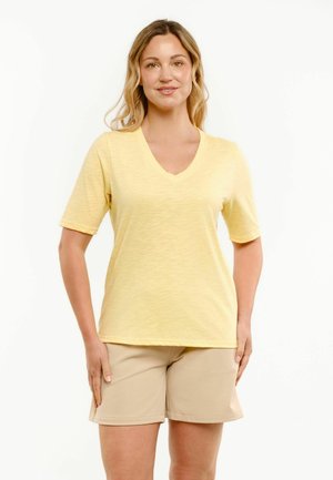 Femme debout portant un t-shirt jaune à manches courtes avec col en V et un short beige longueur genou sur fond blanc uni.