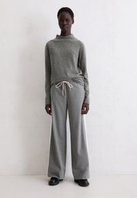 Grauer Strickpullover mit hohem Kragen, kombiniert mit grauen Hosen mit weitem Bein und weißem Seitenstreifen. Glatte Textur, legeres Design.