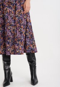 Sukně s květinovým vzorem v purpurové, oranžové a bílé na tmavém pozadí, doplněná černými koženými nadkolenkovými botami na robustním podpatku.