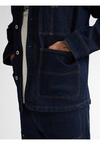 Mørkeblå denimjakke med to forlommer, knaplukning og kontraststing. Vist sammen med matchende denimbukser.