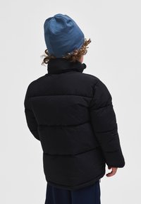 Enfant aux cheveux bouclés portant un bonnet bleu et une doudoune noire, vu de dos sur un fond clair uni.