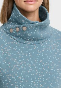 Blusa suave de color azul con un cuello alto y doblado que cuenta con tres botones decorativos. La tela tiene un patrón de pequeños círculos en azul, rosa y blanco.