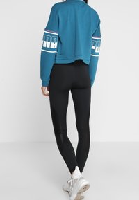 Teal kortärmad sweatshirt med vita och lila ränder på ärmarna, kombinerad med svarta leggings och vita sneakers, med en mjuk textur.