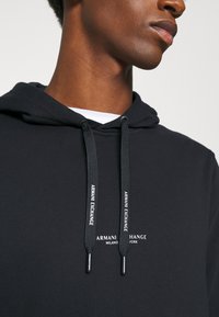 Sudadera negra con cordones que presenta el logo de "Armani Exchange" en blanco. Tejido suave y corte clásico con textura acanalada alrededor de la capucha.