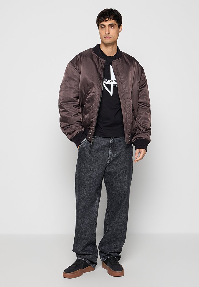 Joven con chaqueta bomber marrón oscuro brillante, camiseta negra con gráfico, vaqueros sueltos gris oscuro y zapatillas negras con suelas marrones.