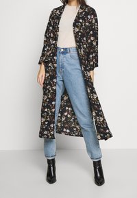 Kvinna iklädd en ljusbeige topp, högmidjade ljusblå jeans, svart blommig lång kimono och svarta spetsiga ankelstövlar mot vit bakgrund.