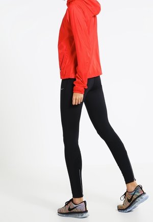 Osoba ubrana w czerwoną kurtkę z kapturem, czarne legginsy i kolorowe buty do biegania marki Nike, stojąca w pozycji bocznej na białym tle.