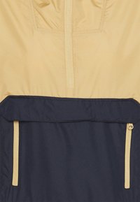 Ljusbeige och marinblå vindjacka med dragkedja framtill och två sidofickor; slät textur med matt finish och kontrasterande färgblock.