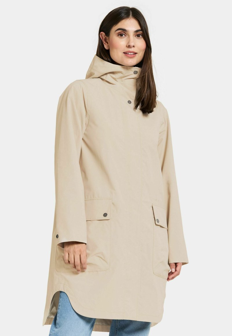 Beige waterdichte jas met capuchon, voorzien van twee voorzakken, drukknoopsluitingen en een rechte zoom. Gladde textuur en een losse pasvorm.