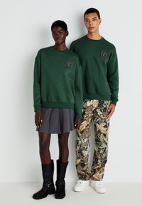 Felpe verdi con logo ricamato, abbinate a una gonna a pieghe grigia, pantaloni mimetici e stivali neri alti fino al ginocchio. Sneakers bianche indossate da un uomo.