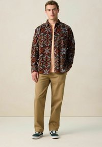 Brown patterned shirt with leaf motifs over a light peach t-shirt, paired with khaki pants and green/white sneakers. Smooth fabric texture.