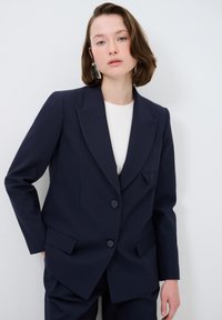 Marinblå skräddarsydd kavaj med dubbelknäppning, slagna lapels och två framfickor, tillverkad av en slät, texturerad tyg.