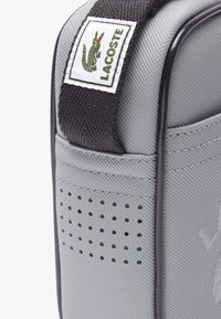 Borsa sintetica grigia con tracolla scura, patch con logo Lacoste, superficie strutturata, design perforato e dettaglio del logo in evidenza sul lato.