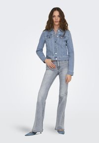 Chaqueta de mezclilla azul claro con un diseño ajustado, cierres de botón y bolsillos delanteros. Combinada con pantalones vaqueros de corte acampanado en azul claro y zapatos de punta azul.