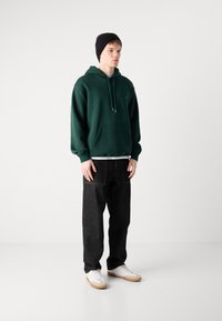 Sudadera verde oscura con un bolsillo canguro, combinada con vaqueros negros holgados y zapatillas blancas con suelas de goma. Gorro negro en la cabeza.
