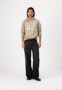 Sudadera beige con el logo "Calvin Klein", combinada con jeans oscuros de pierna ancha y zapatos negros. Tejido suave y corte relajado. Diseño minimalista.