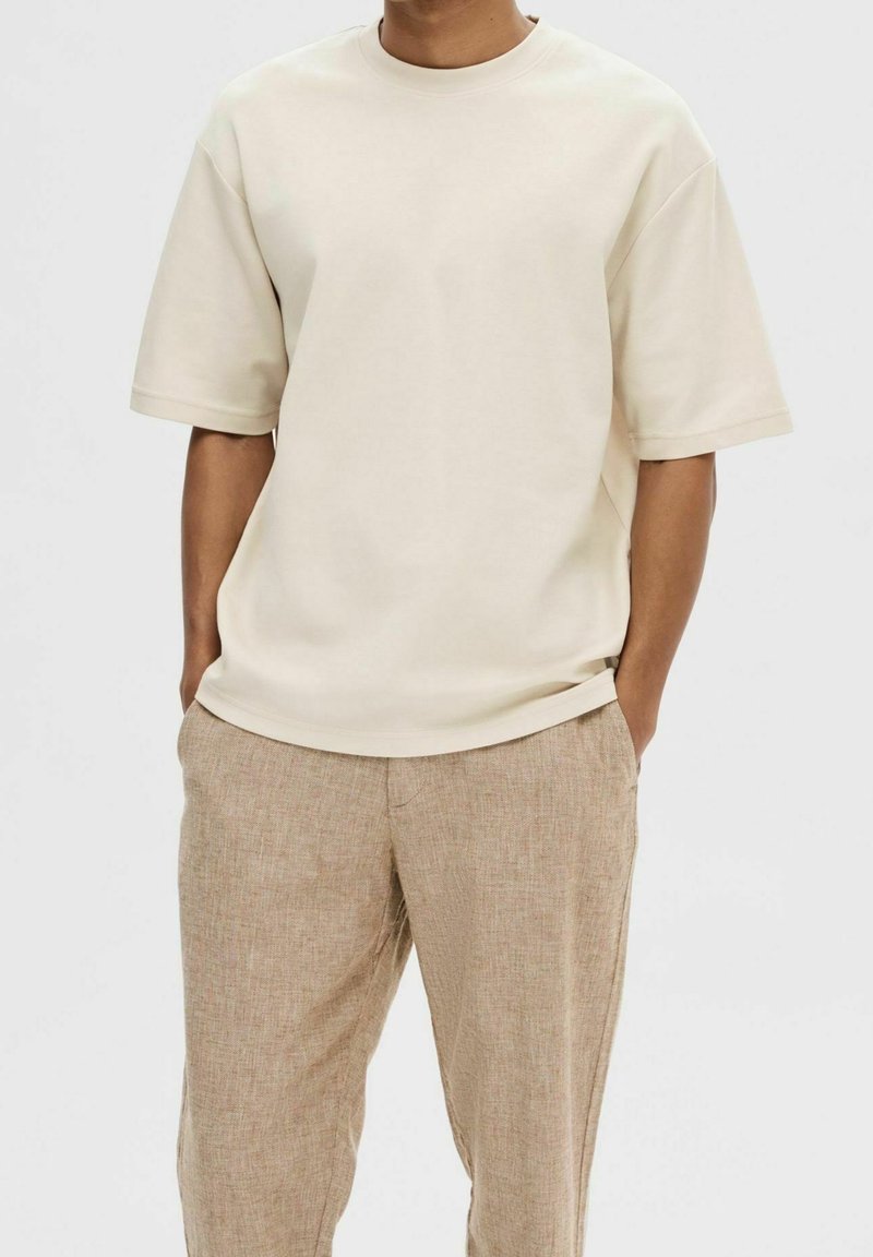 Homme portant un t-shirt beige uni à manches courtes et un pantalon marron clair texturé, les mains dans les poches, sur un fond uni.