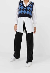 Granatowy sweter w krótki rękaw z niebieskimi diamentowymi wzorami noszony na białej koszuli z guzikami, w zestawie z luźnymi czarnymi spodniami z kordurowej tkaniny i białymi trampkami.