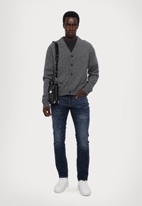 Graue Knopf-Cardigan über einem schwarzen Shirt, kombiniert mit blauen Jeans und weißen Sneakern. Eine schwarze Umhängetasche hängt von der Schulter.