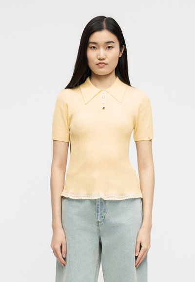 Νεαρή γυναίκα με ίσια σκούρα μαλλιά, ντυμένη με ανοιχτό κίτρινο πλεκτό τοπ με κοντά μανίκια και γιακά, και ανοιχτό μπλε τζιν παντελόνι.