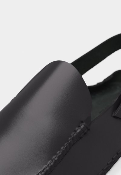 Gros plan sur une chaussure noire en cuir à enfiler avec des coutures détaillées et une sangle élastique noire au talon.