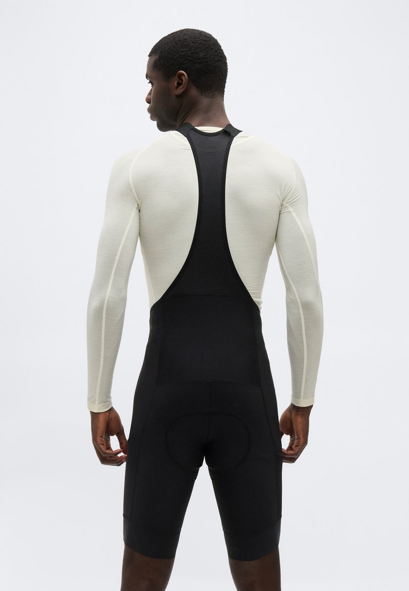 Man wearing white long-sleeve shirt and black cycling bib shorts, shown from the back against a plain light background.