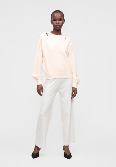 Femme portant un sweat-shirt beige clair avec des découpes aux épaules, un pantalon blanc à jambes larges et des talons noirs à bouts pointus, debout devant un fond uni.