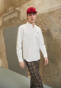 Chemise blanche à boutons avec col, détail de logo noir sur la poitrine, associée à un pantalon carreauté marron. Le modèle porte une casquette rouge. L'arrière-plan présente un mur texturé.