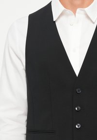 Črn službeni suknjič z gladko teksturo, z V-izrezom, petimi gumbi in stransko žepom. Nosi se čez belo srajco s kragnjo.