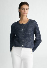 Navy gebreide cardigan met ronde hals, lange mouwen, textuurafwerking, knopen aan de voorkant en twee opgestikte zakken. Gekaapt met witte broek.