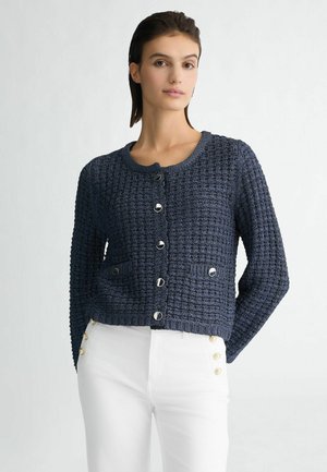 Cardigan lavorato a maglia blu navy con scollatura rotonda, maniche lunghe, finitura testurizzata, bottoni davanti e due tasche a toppa. Indossato con pantaloni bianchi.
