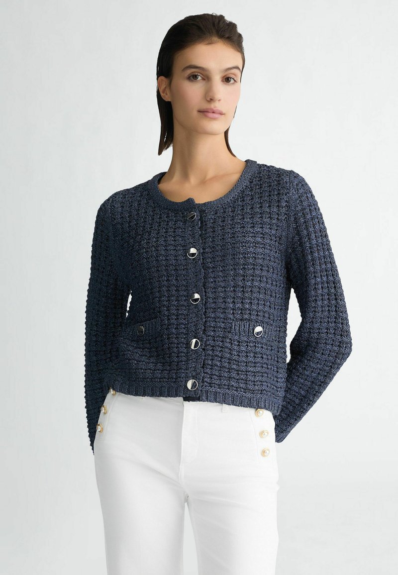 Navy gebreide cardigan met ronde hals, lange mouwen, textuurafwerking, knopen aan de voorkant en twee opgestikte zakken. Gekaapt met witte broek.
