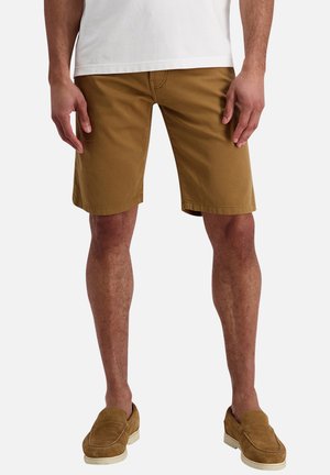 Mand iført brune knælange shorts, lysebrune ruskind slip-on sko og en hvid skjorte, stående med hænderne afslappet ved siden.
