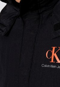 Svart jacka med en slät textur, med en överdimensionerad krage, kardborrefästen och en orange "cK" logotyp framtill.