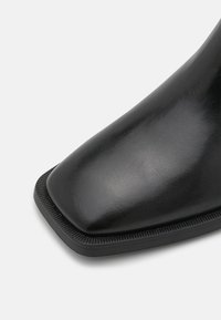Botín de cuero negro con punta cuadrada, textura suave y suela de goma contrastante con un borde texturizado a lo largo del borde.