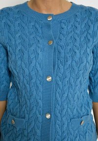 Blå stickad cardigan med texturerat kabelmönster, korta ärmar och två framfickor, med silverfärgade knappar och ribbad halsringning.