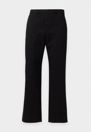 Homme portant un polo noir à manches courtes à motifs, pantalon noir, baskets blanches, debout avec une main dans la poche.