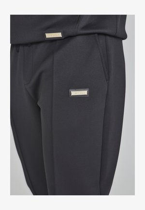 Pantalon noir en tissu lisse avec une coupe ajustée. Il présente des poches latérales et une petite étiquette de marque dorée sur la taille.