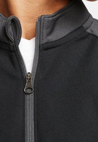 Svart zip-up-jacka med grå detaljer, med en hög krage och slät tygtextur. Inkluderar en metallic dragkedja framtill.