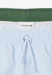 Svetlomodré plavky s elastickým pásom, zeleným akcentom a bielou sieťovinou. Obsahujú šnúrku a štítok s logom Lacoste.