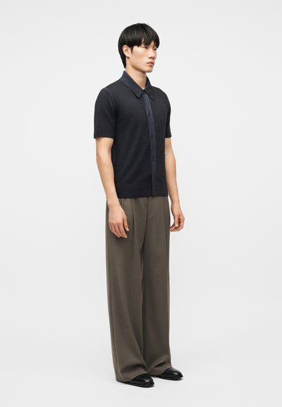 Νεαρός άνδρας που στέκεται, φορώντας ένα μαύρο πουκάμισο με κοντά μανίκια και κουμπιά, πλατύ γκρι παντελόνι και μαύρα παπούτσια, μπροστά σε απλό φωτεινό φόντο.