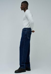 Jeans de mezclilla azul oscuro con corte recto, combinados con una camisa blanca de manga larga, que cuentan con bolsillos traseros y detalles minimalistas en el hardware.