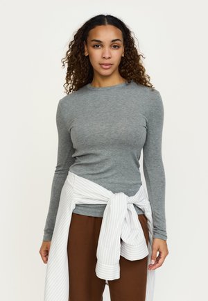 Grijs T-shirt met lange mouwen en ronde hals, gecombineerd met een bruine broek en een wit-grijs gestreept overhemd dat rond de taille is gestrikt.