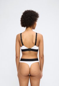 Γυναίκα με σγουρά μαλλιά, εμφανιζόμενη από πίσω, φοράει λευκό και μαύρο σουτιέν και ταιριαστό στρινγκ εσώρουχο,  μπροστά σε απλό φόντο.
